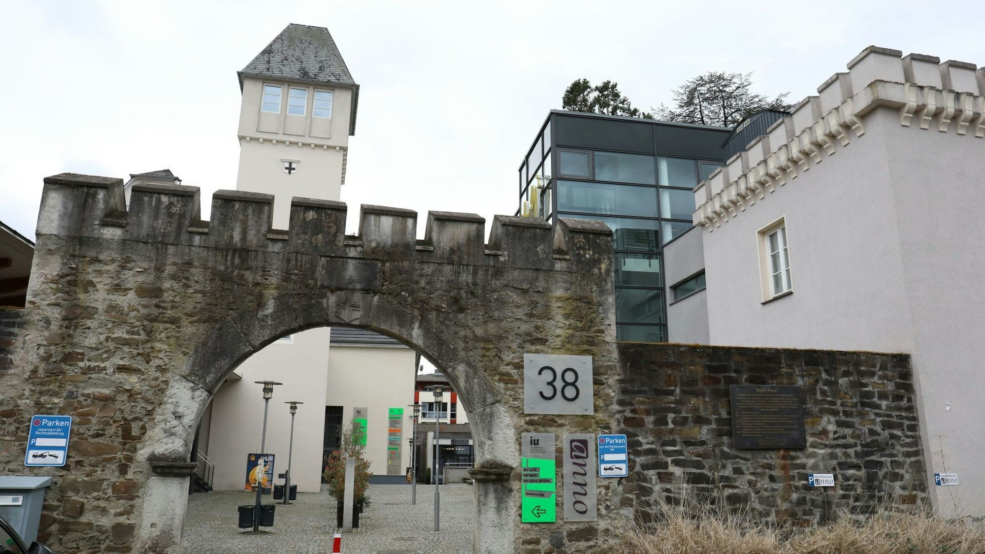Hinter einer mittelalterlich anmutenden Mauer mit Torbogen erhebt sich ein Turm, daneben steht ein gläsernes Gebäude.