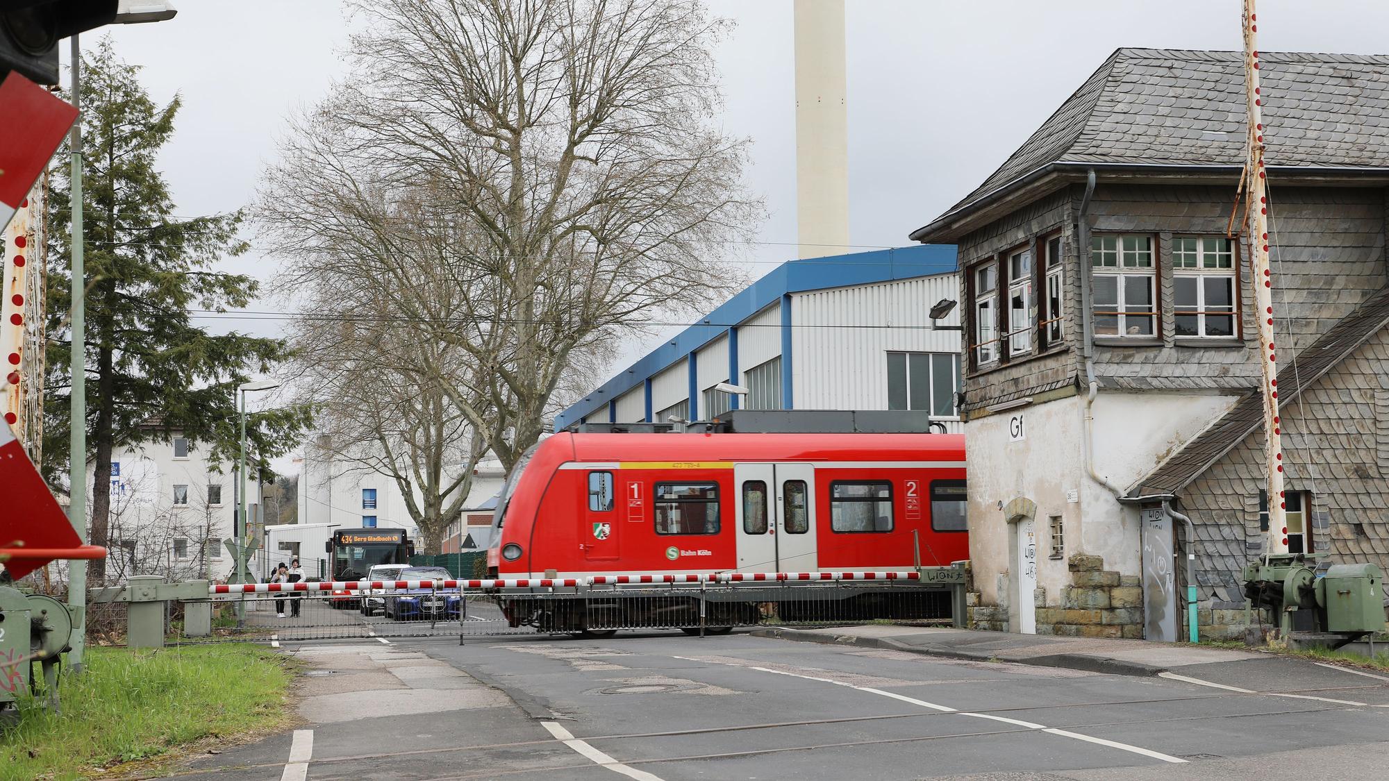 Zwischen den geschlossenen Schranken des Bahnübergangs an der Tannenbergstraße in Bergisch Gladbach fährt eine S-Bahn hindurch. Rechts ist ein historisches Stellwerk zu sehen, davor eine weitere, nicht geschlossene Schrankenanlage. Im Hintergrund warten Autos und ein Bus vor den geschlossenen Schranken.