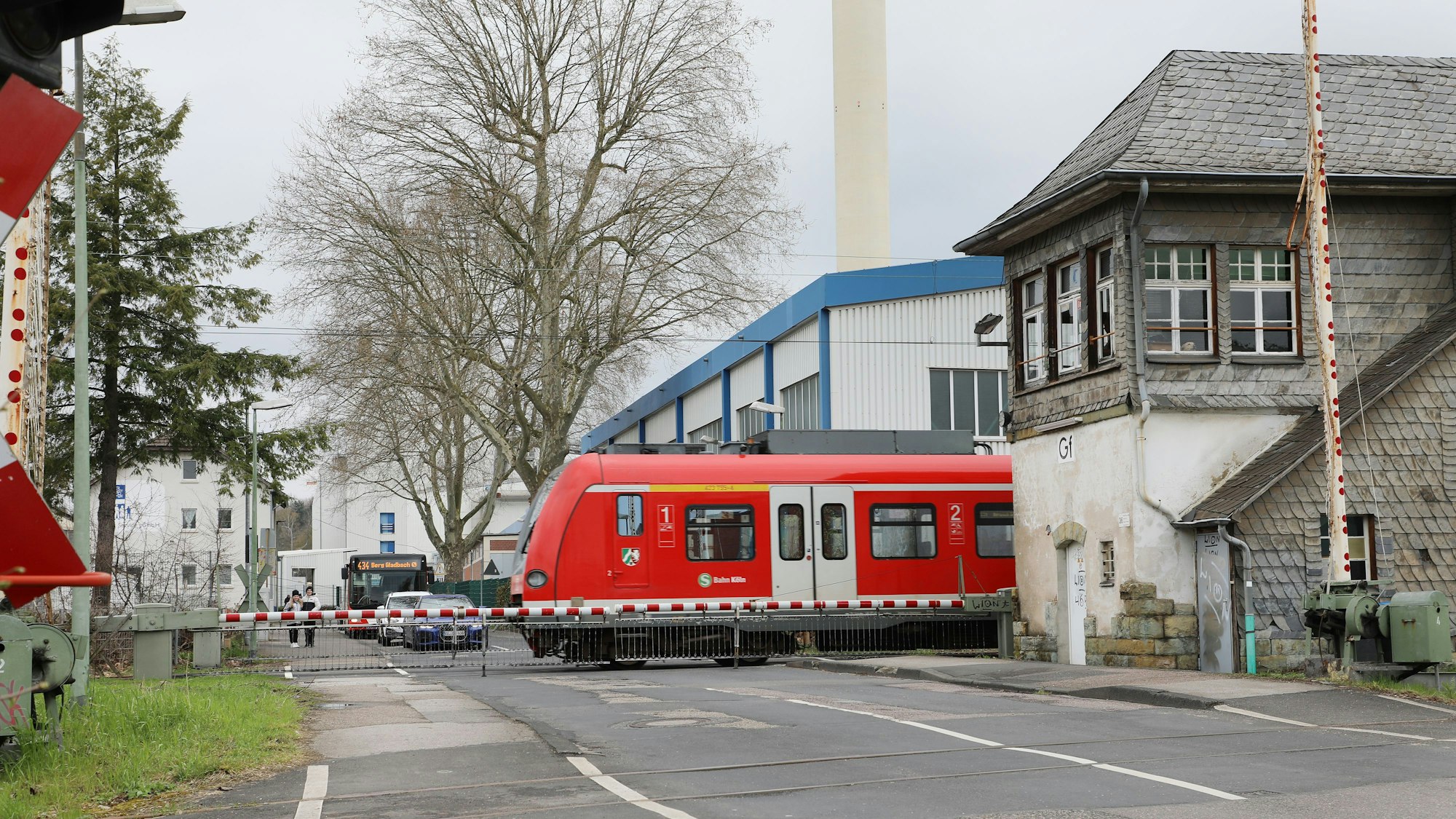 Zwischen den geschlossenen Schranken des Bahnübergangs an der Tannenbergstraße in Bergisch Gladbach fährt eine S-Bahn hindurch. Rechts ist ein historisches Stellwerk zu sehen, davor eine weitere, nicht geschlossene Schrankenanlage. Im Hintergrund warten Autos und ein Bus vor den geschlossenen Schranken.