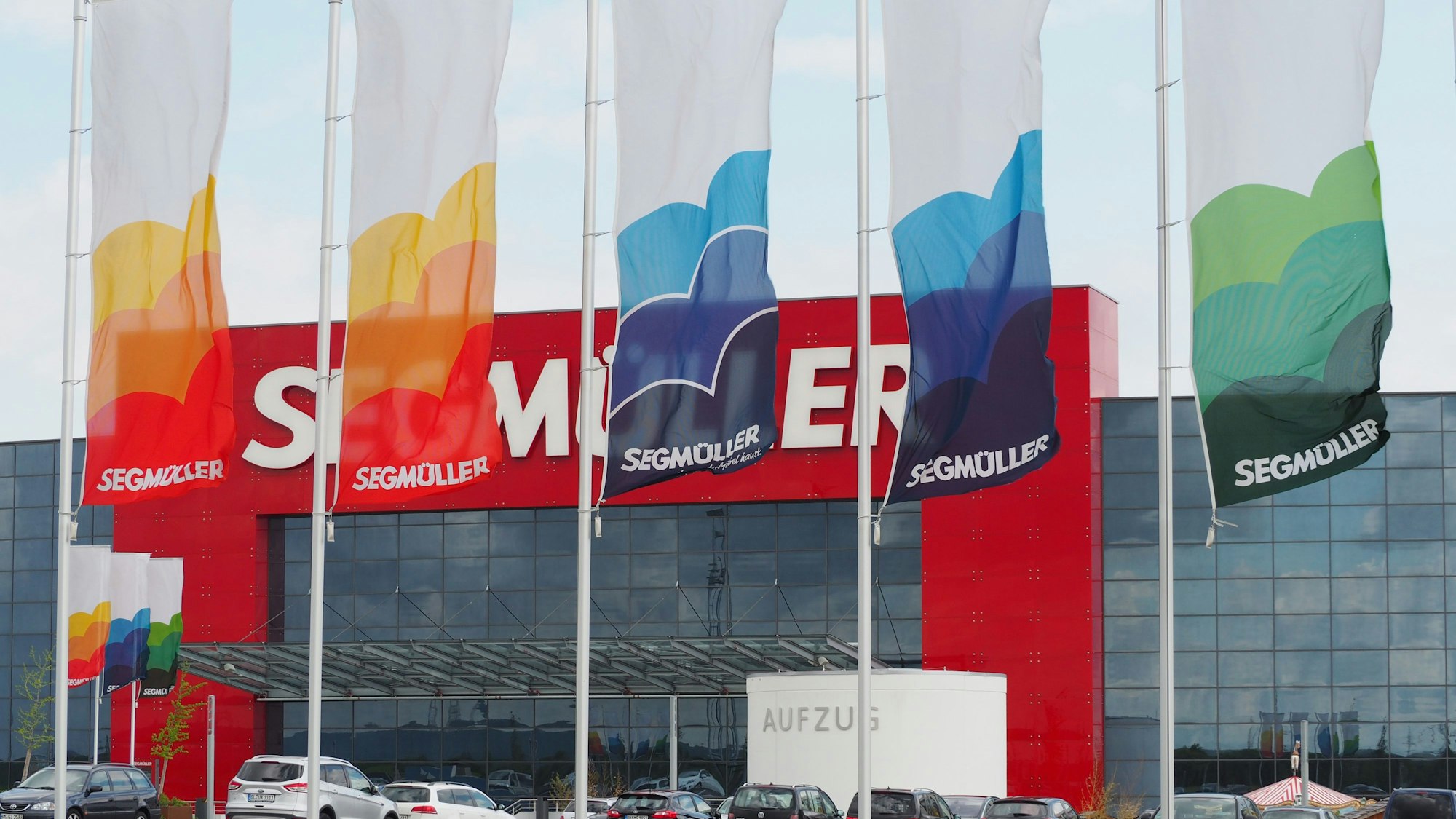Im Vordergrund fünf Fahnen, im Hintergrund geparkte Fahrzeuge und die markante Fassade eines Einrichtungshauses.