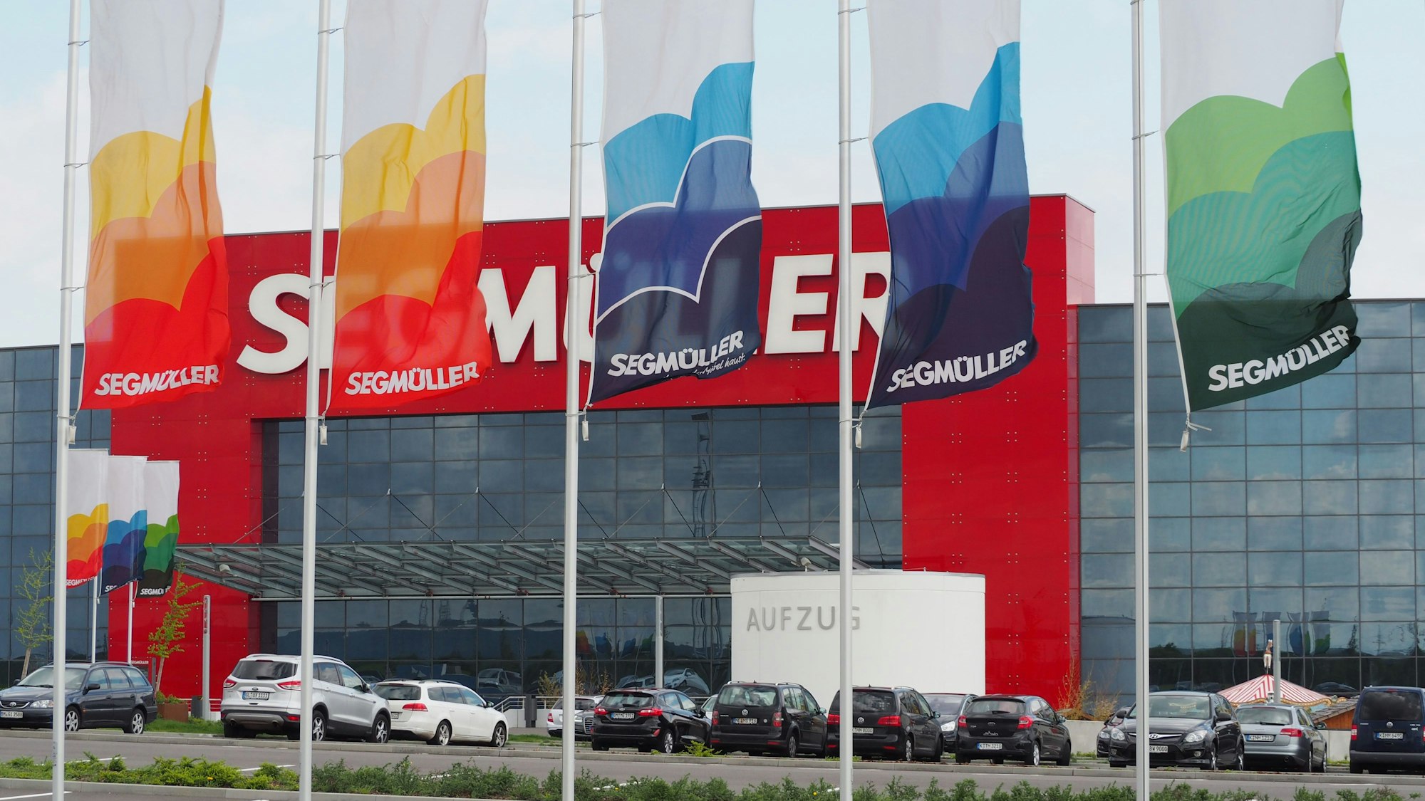 Das Foto zeigt den Eingang und den Parkplatz des Einrichtungshauses Segmüller. Davor hängen bunte Fahnen.