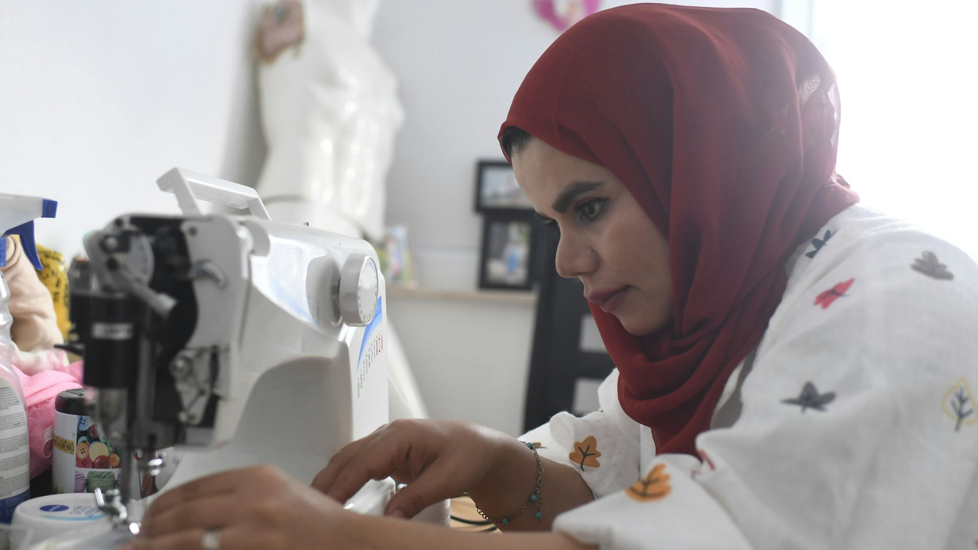 Nahida Mohammadi sitzt an ihrer Nähmaschine und näht. Sie trägt eine weiße Bluse mit buntem Muster und ein dunkelrotes Kopftuch.