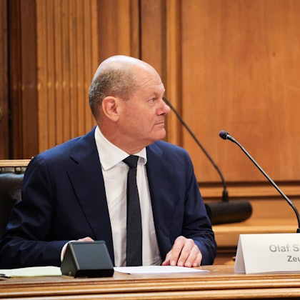 Bundeskanzler Olaf Scholz (SPD) sitzt auf seinem Platz auf der Senatsbank mit einem Plastikschild mit dem Text ·Olaf Scholz Zeuge· vor Beginn einer Sitzung des Parlamentarischen Untersuchungsausschusses ·Cum-Ex· im Plenarsaal der Hamburgischen Bürgerschaft im Rathaus.