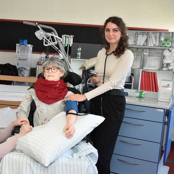 Zwei junge Frauen stehen in einem Krankenzimmer. In ihrer Mitte ist eine Puppe mit dem Aussehen einer älteren Frau, an der sie den Patientenkontakt üben.