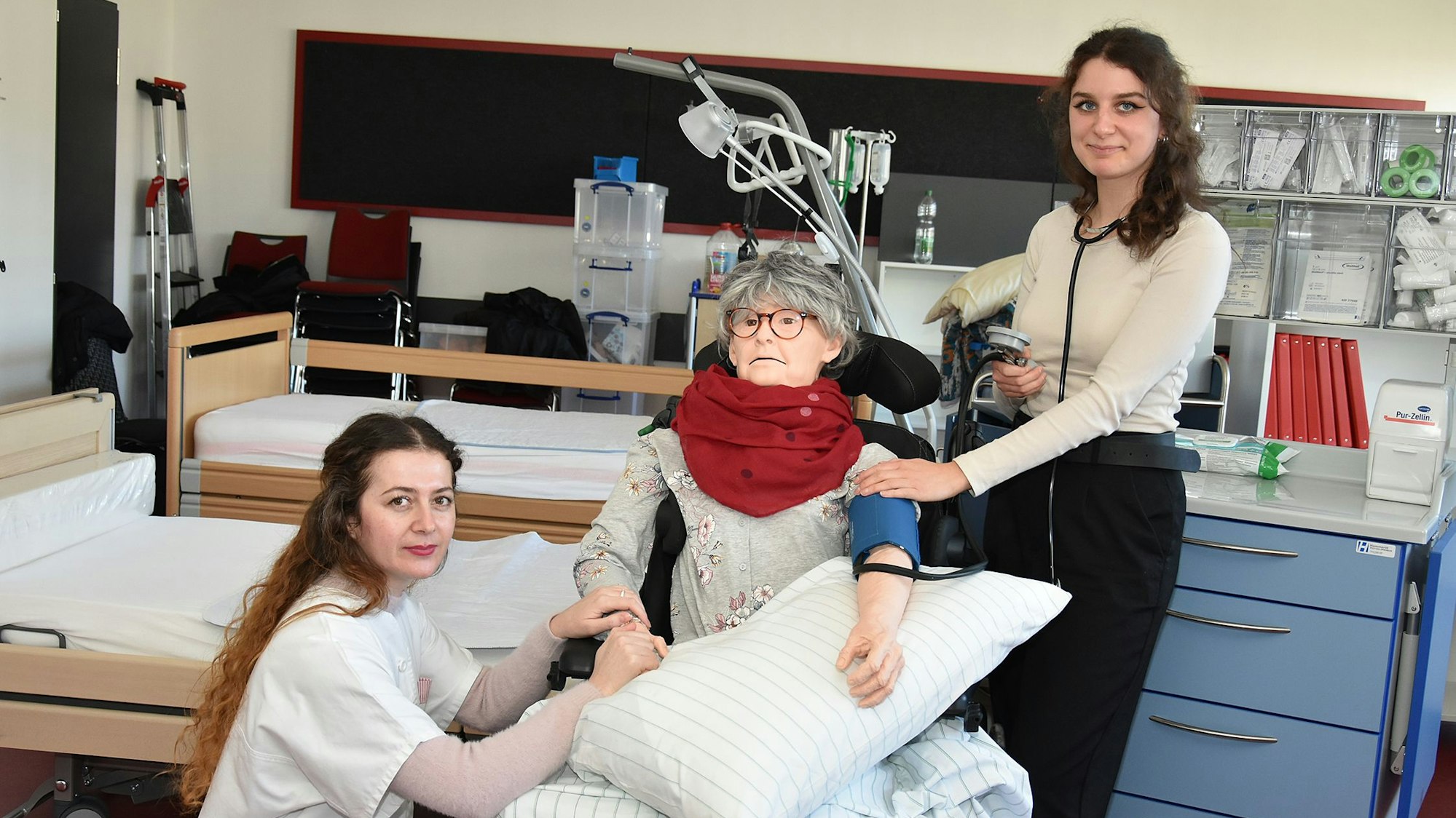 Zwei junge Frauen stehen in einem Krankenzimmer. In ihrer Mitte ist eine Puppe mit dem Aussehen einer älteren Frau, an der sie den Patientenkontakt üben.