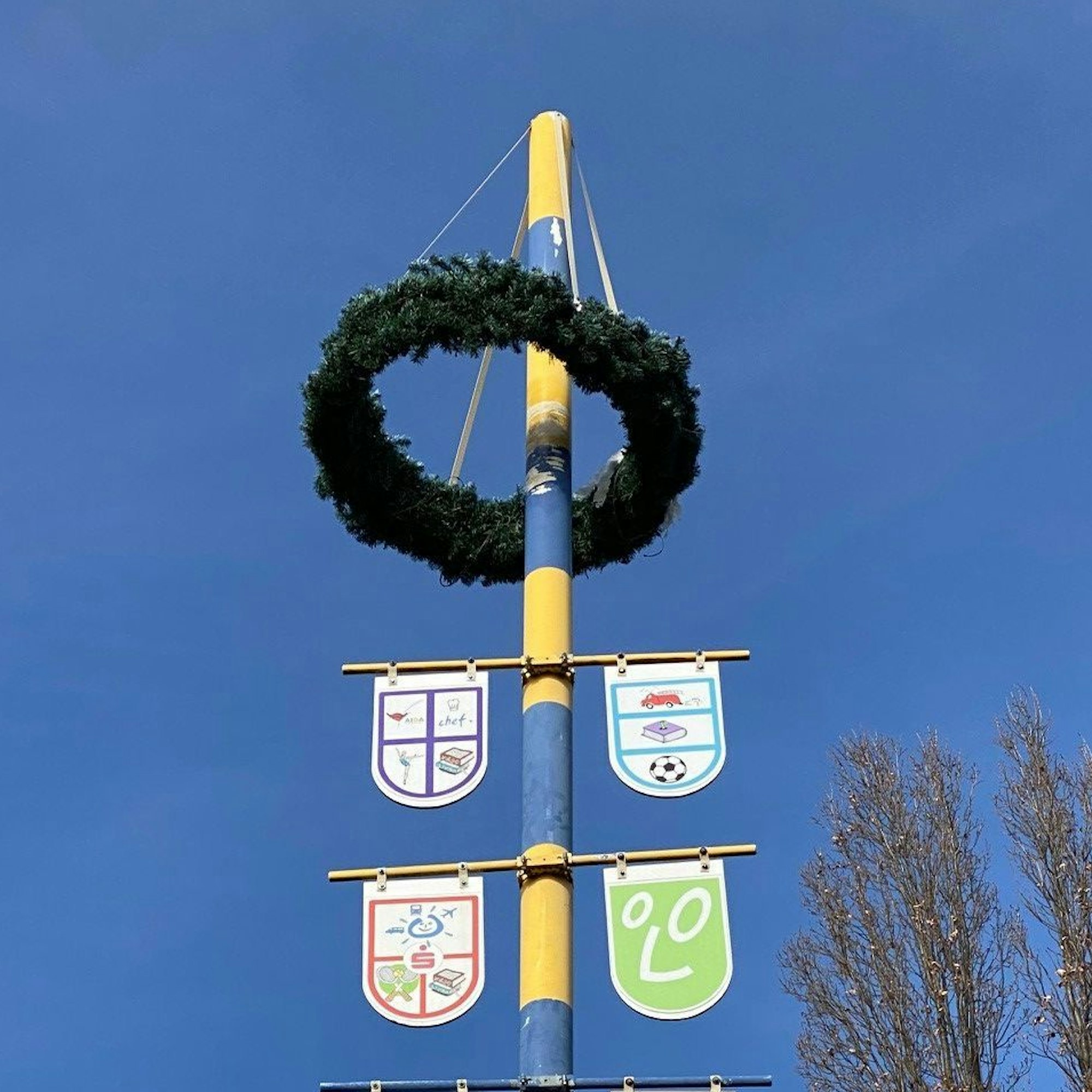 Blau-goldener Maibaum-Mast, geschmückt mit Kranz und Wappen