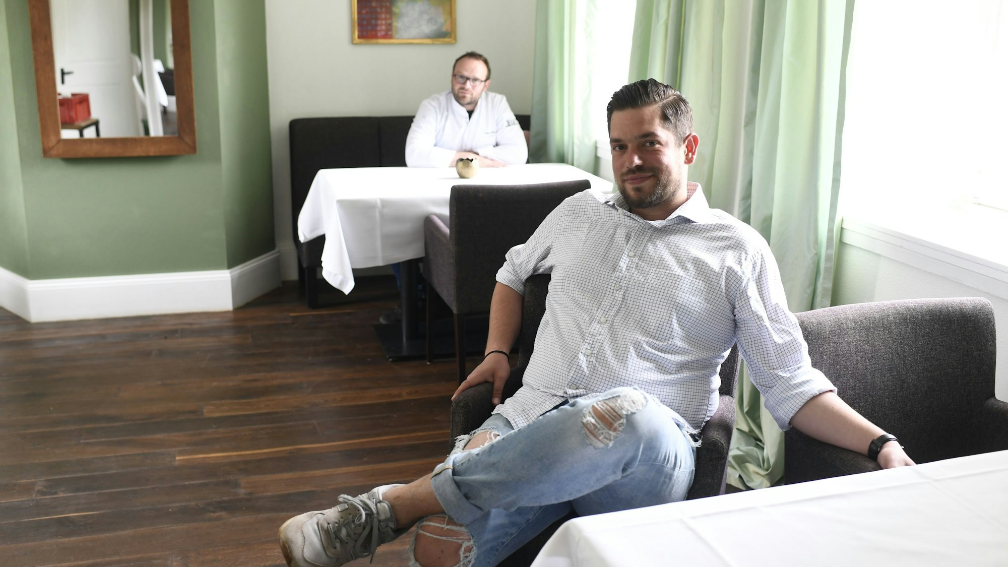 Im Vordergrund sitzt Filip Czmok, Küchenchef im Bembergs Häuschen, im Hintergrund Oliver Röder, ehemaliger Küchenchef und Küchendirektor auf Burg Flamersheim, an Tischen in dem in Grüntönen dekorierten Restaurant.