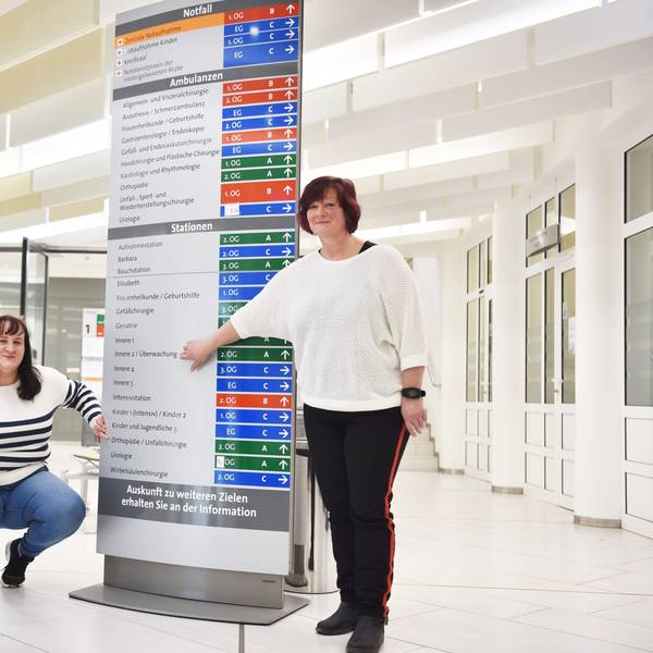Zwei Frauen zeigen auf einen Wegweiser, der zu den Stationen in einem Krankenhaus führt.