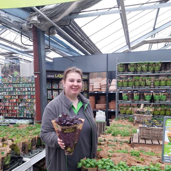 Eine Gartenexpertin in einem Blumenladen hält grinsend einen Blumentopf vor sich.