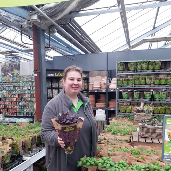 Eine Gartenexpertin in einem Blumenladen hält grinsend einen Blumentopf vor sich.