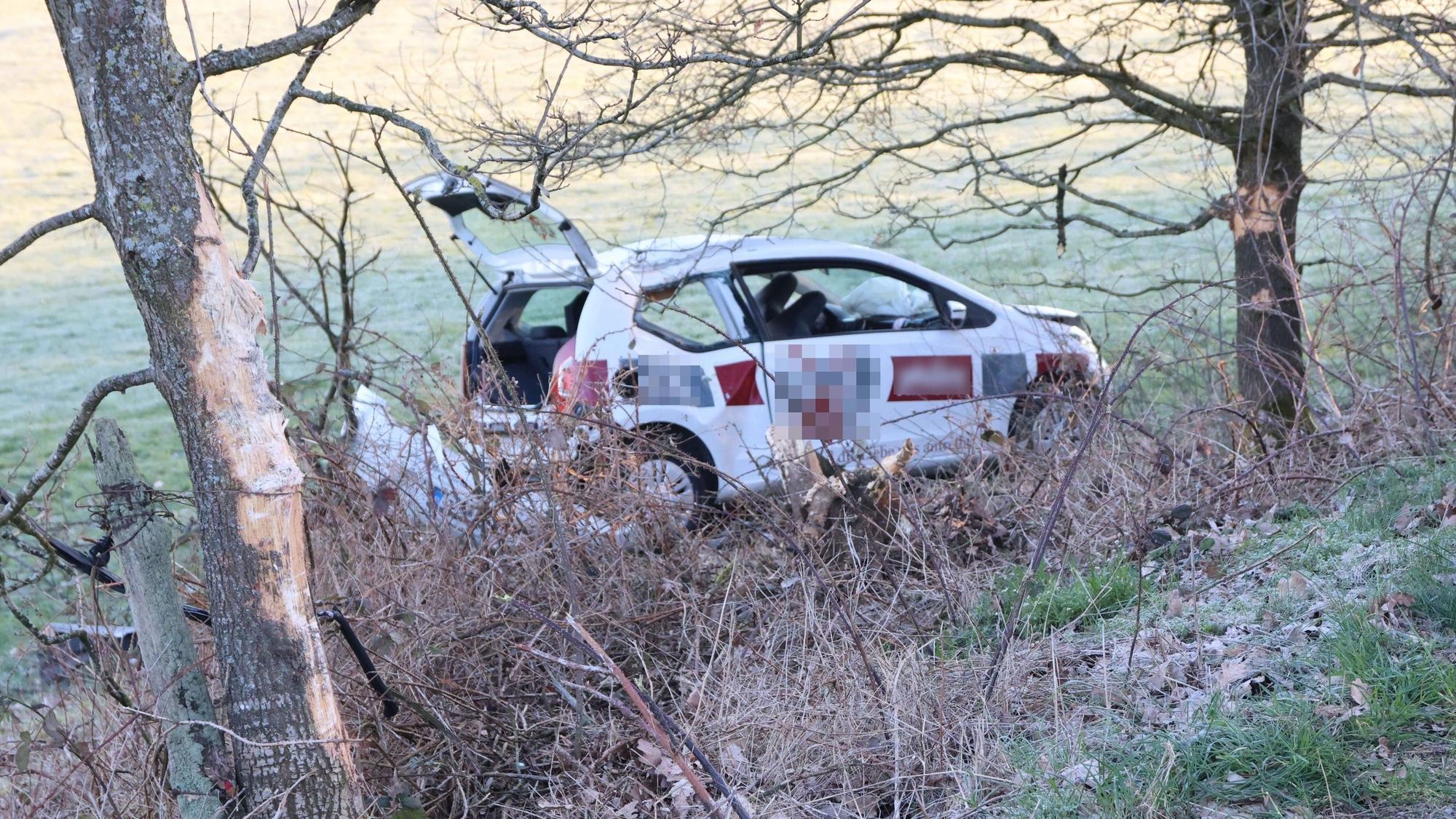 Ein Auto steht demoliert in einem Hang abseits der Straße zwischen zwei Bäumen.