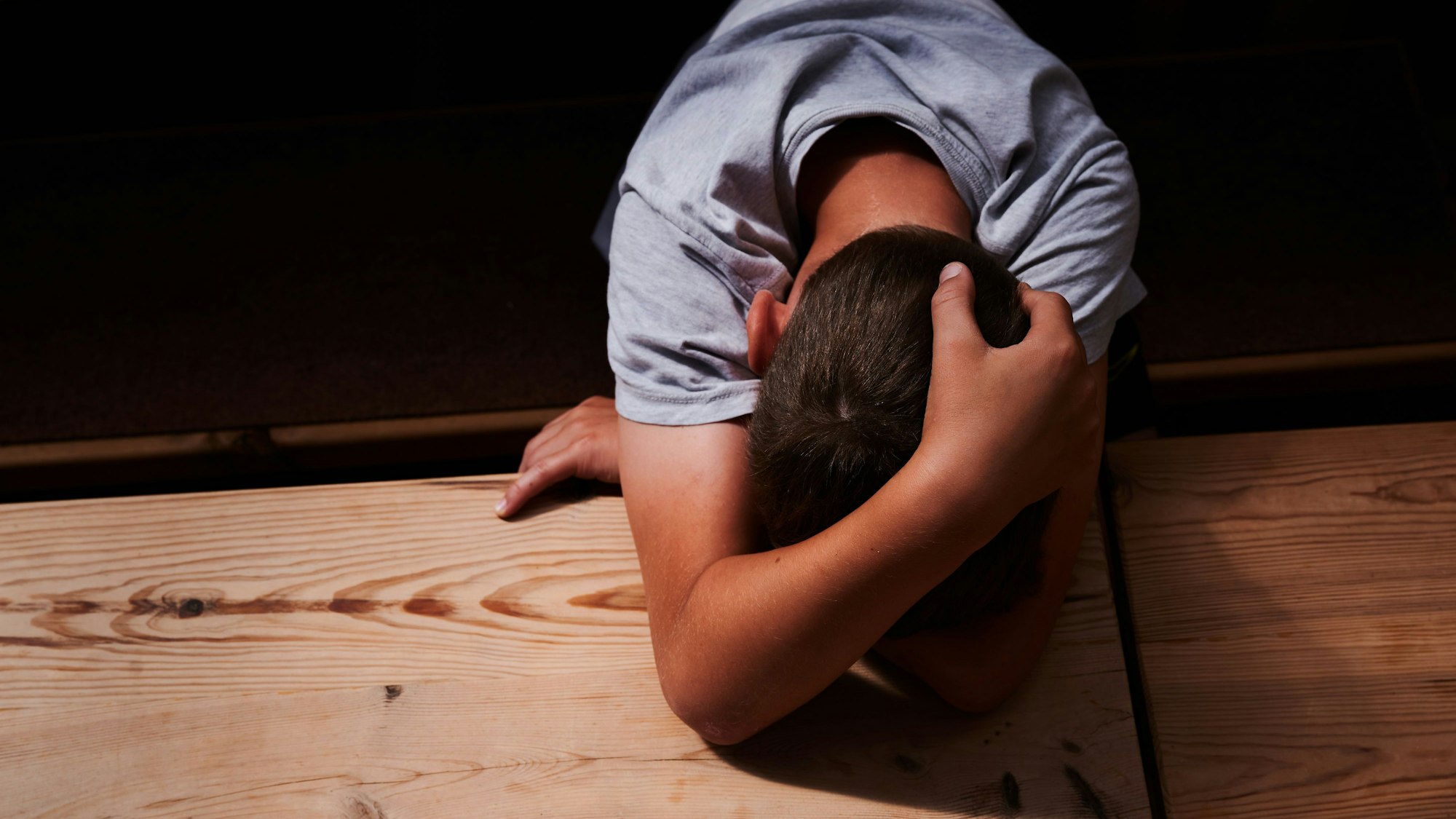 Ein Kind versteckt sein Gesicht unter seinen Armen und liegt mit dem Kopf auf einem Tisch.