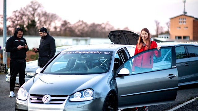 Michelle Engel steht an ihrem Golf R32. Im Hintergrund schauen zwei Männer auf ihr Handy.