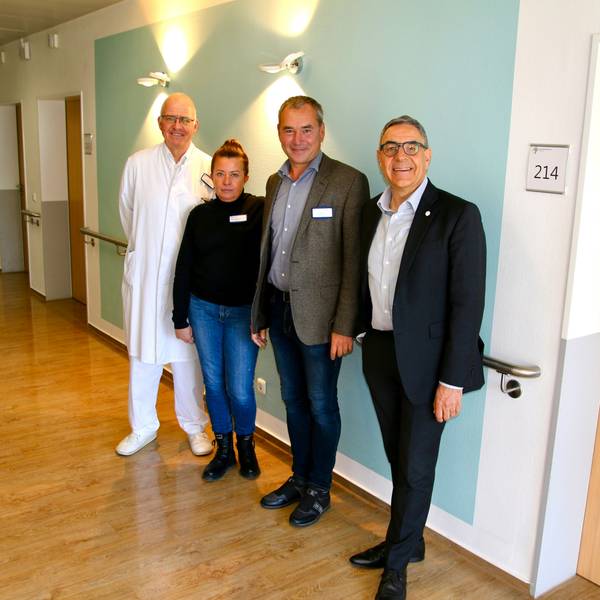 Drei Männer und eine Frau posieren lächelnd für eine Bildaufnahme vor einer Wand im Krankenhaus. Links im Hintergrund sind Türen zu sehen, die zu Patientenzimmern führen. Rechts oben im Bild ist ein Schild mit der Aufschrift "214" zu sehen.