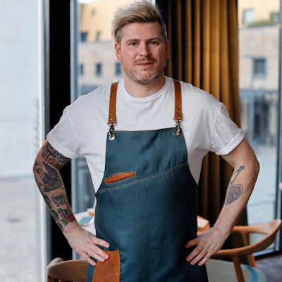 Daniel Gottschlich, Küchenchef und Inhaber des Ox & Klee im Kölner Rheinauhafen psoiert mit einer modern gestalteten Kochschürze für die Kamera.