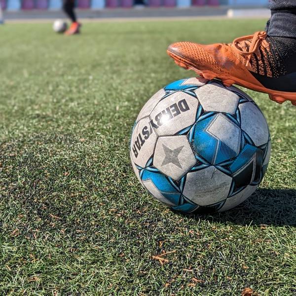 Das Foto zeigt einen Fuß mit Fußballschuh, der auf einem Ball auf einem Kunstrasenplatz steht.