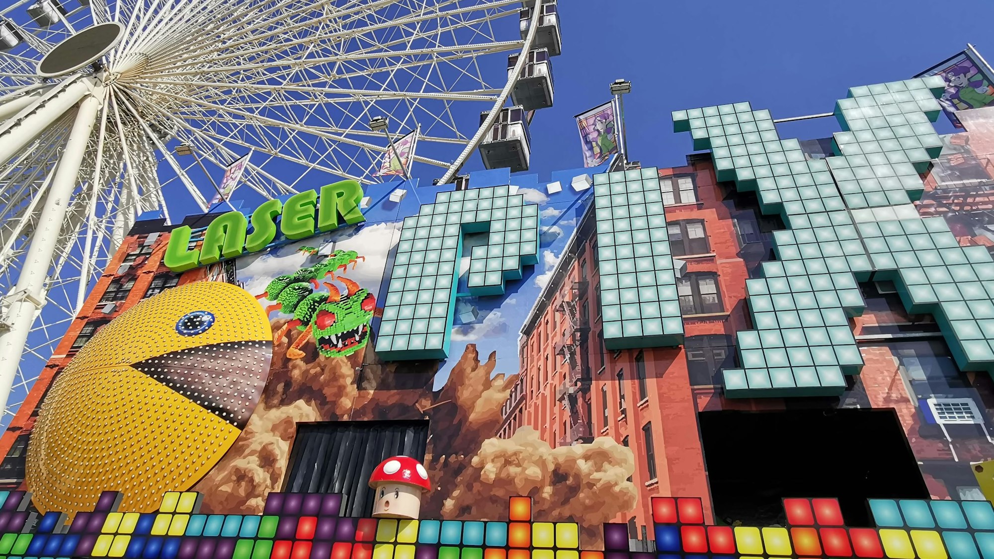 Premiere in Köln: Beim „Laser Pix“ fährt man wie durch eine Geisterbahn, zielt dabei aber mit Laserpistolen auf Pixel bekannter Figuren. Die kunterbunte Fassade des großen Fahrgeschäftes beseht zumeist aus bunten Pixelquadraten, darüber ragt das weiße Riesenrad in den blauen Himmel.