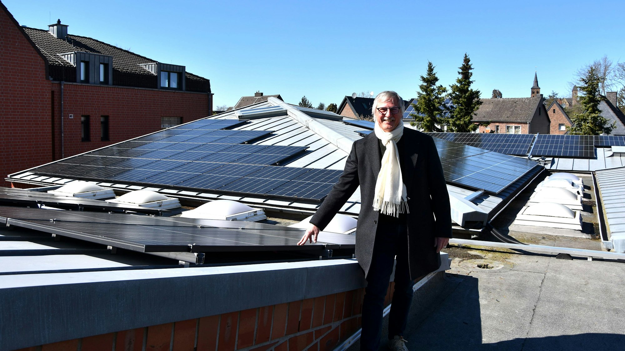 Ein grauhaariger Mann steht auf einem Hausdach. Der Mann trägt Brille, Schal und Mantel. Mit der rechten Hand lehnt der Mann mit gespreizten Fingern an einer Photovoltaikanlage.
