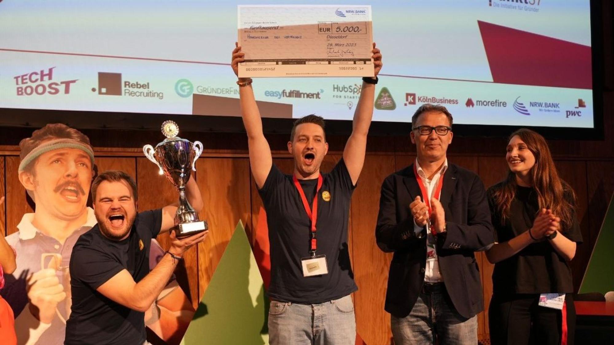 Mathias (l.) hält den Pokal in den Händen, Daniel zeigt den Check. Daneben applaudieren ein Mann und eine Frau.