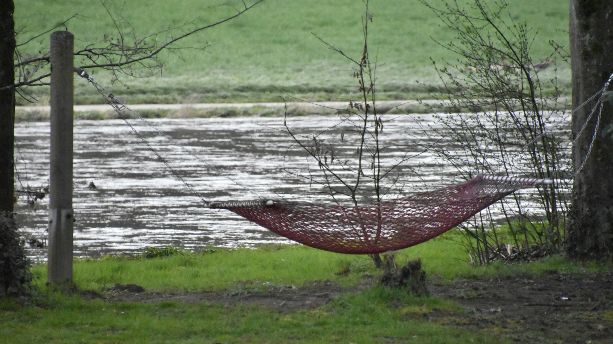 Eine rote Hängematte ist gespannt. Im Hintergrund sieht man Wasser und grüne Wiesen.