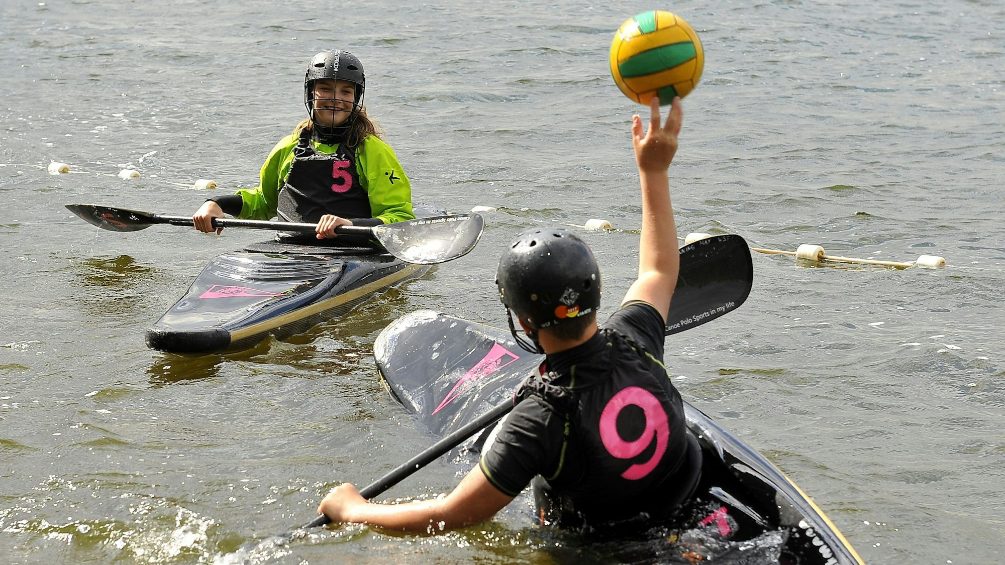 Zwei Kanupolo-Spielende der Schülerklasse sitzen rudernd in ihren Einer-Kanus. Der vorne im Bild befindliche Kanupolospieler wirft einen Ball zu einer Kanupolospielerin. Um die beiden herum ist Wasser.