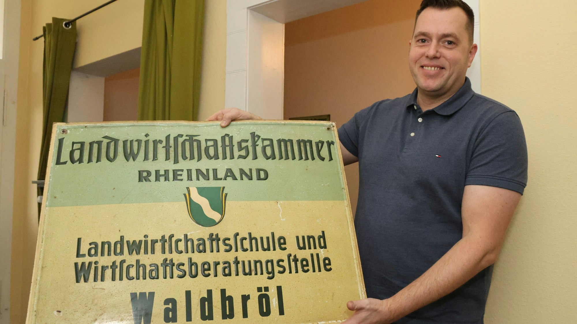 David Hahn hält ein altes Blechschild in der Hand. Darauf steht: Landwirtschaftsklammer Rheinland – Landwirtschaftsschule und Wirtschaftsberatungsstelle Waldbröl.