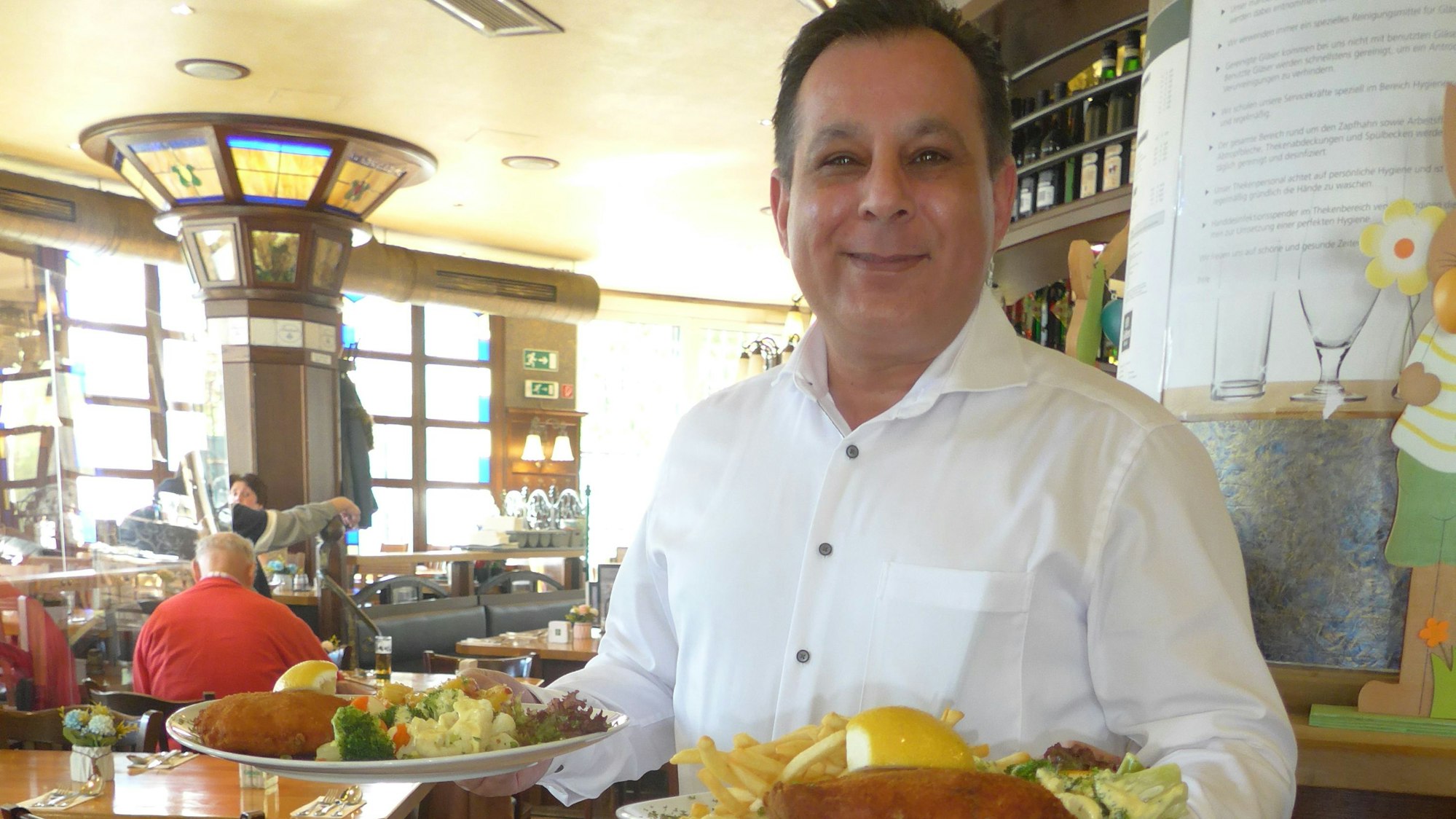 Iakovos Sidiropoulos, Geschäftsführer des „Gilden Brauhaus“ in Köln-Mülheim, steht in seinem Restaurant und hat zwei Teller mit Speisen in den Händen.