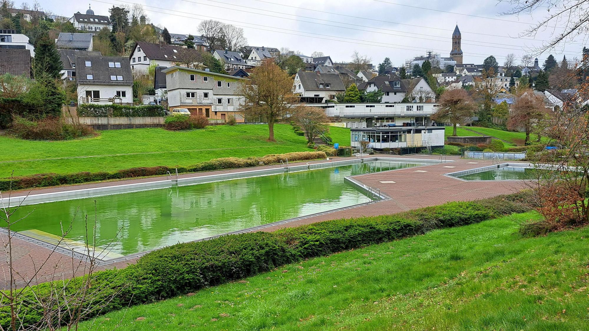 Im Vordergrund liegt ein Frebad, dessen Wasser grün vor Algen ist. Im Hintergrund sind Häuser, am Horizont eine Kirche zu sehen.