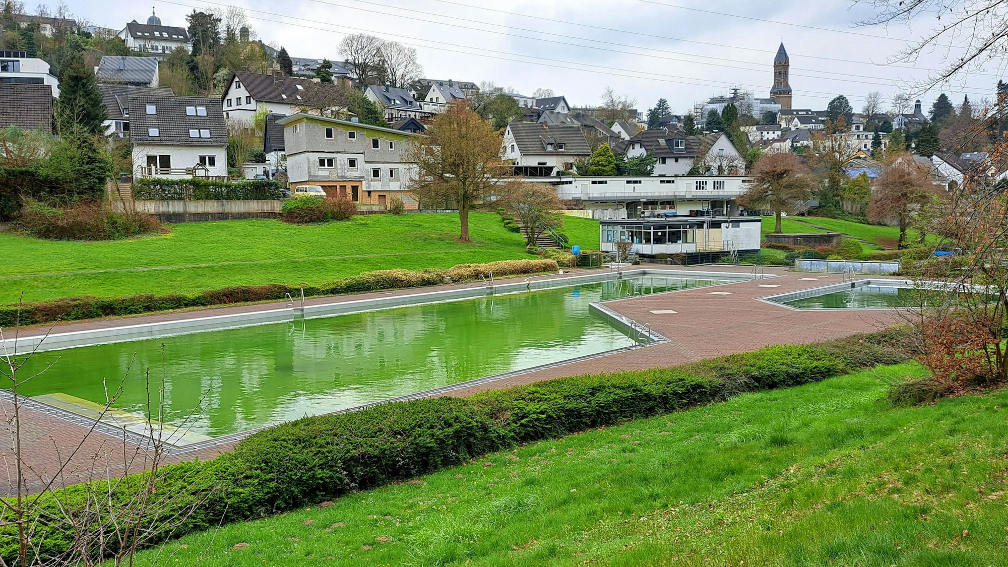 Im Vordergrund liegt ein Frebad, dessen Wasser grün vor Algen ist. Im Hintergrund sind Häuser, am Horizont eine Kirche zu sehen.