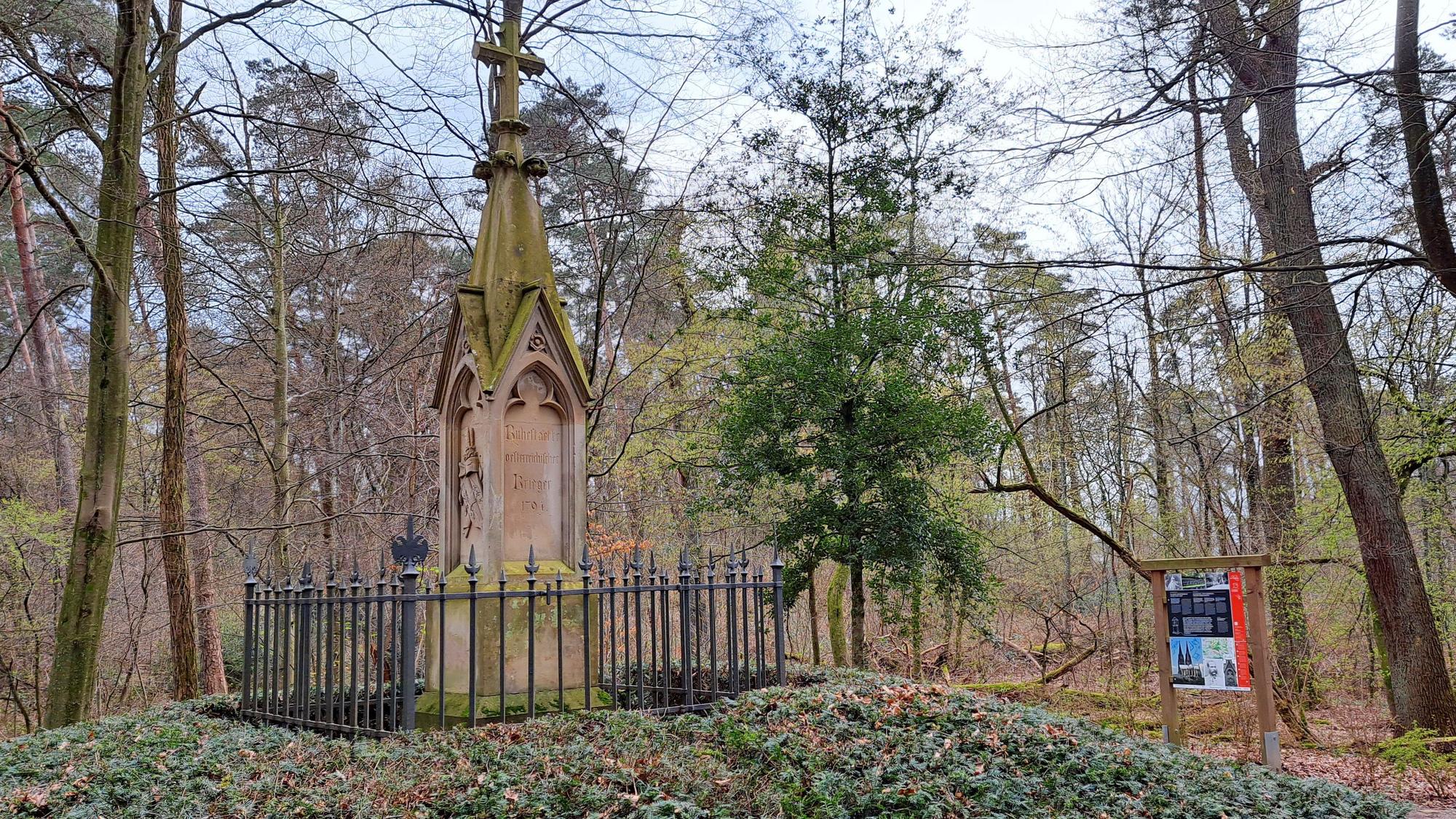 Ein gotisches Denkmal steht auf einem Hügel. Rechts im Hintergrund ist eine bunter Informationstafel zu sehen.