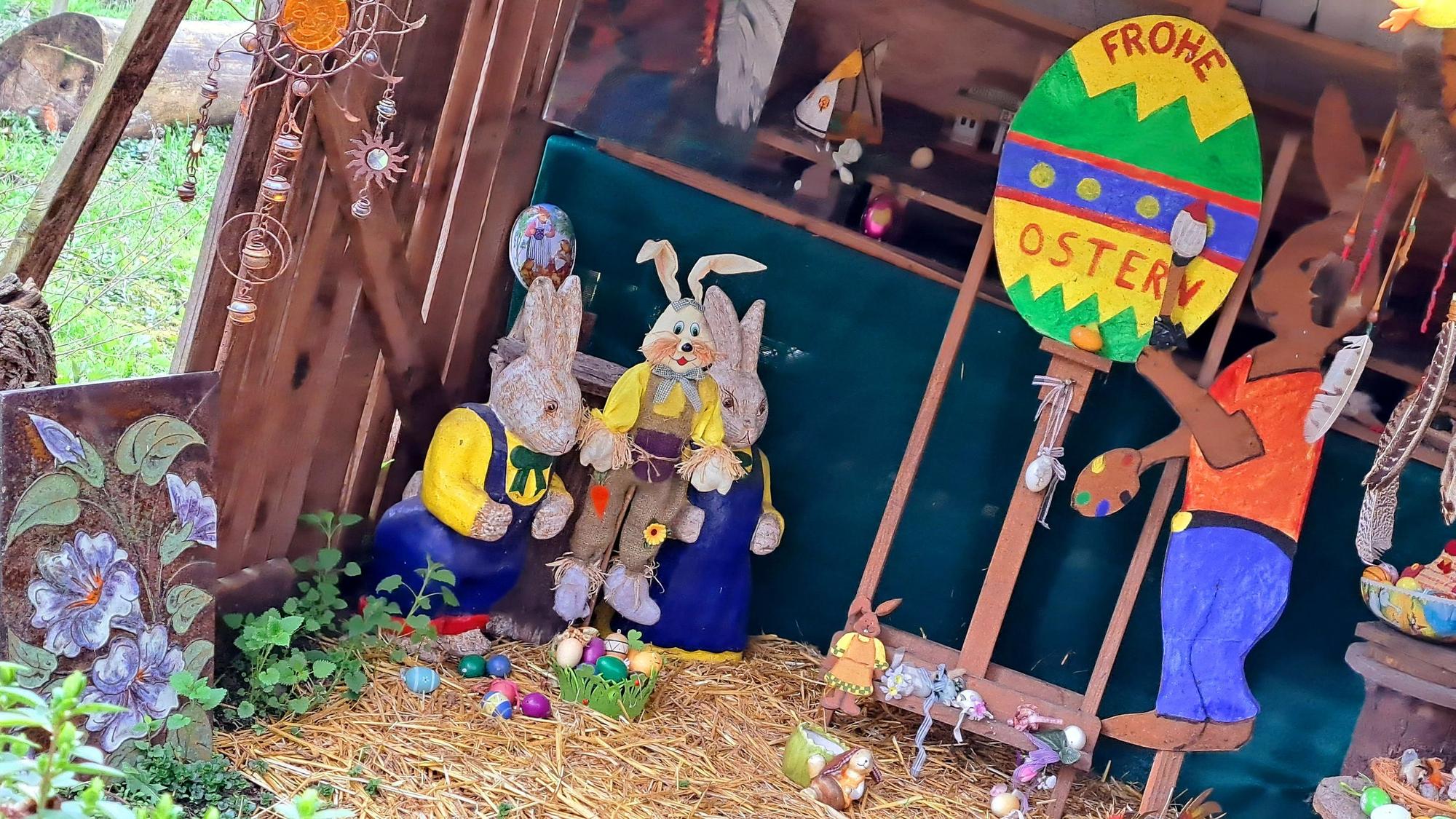 Hasenfiguren stehen in einem Unterstand, auf einem großen bunten Osterei steht „Frohe Ostern“.