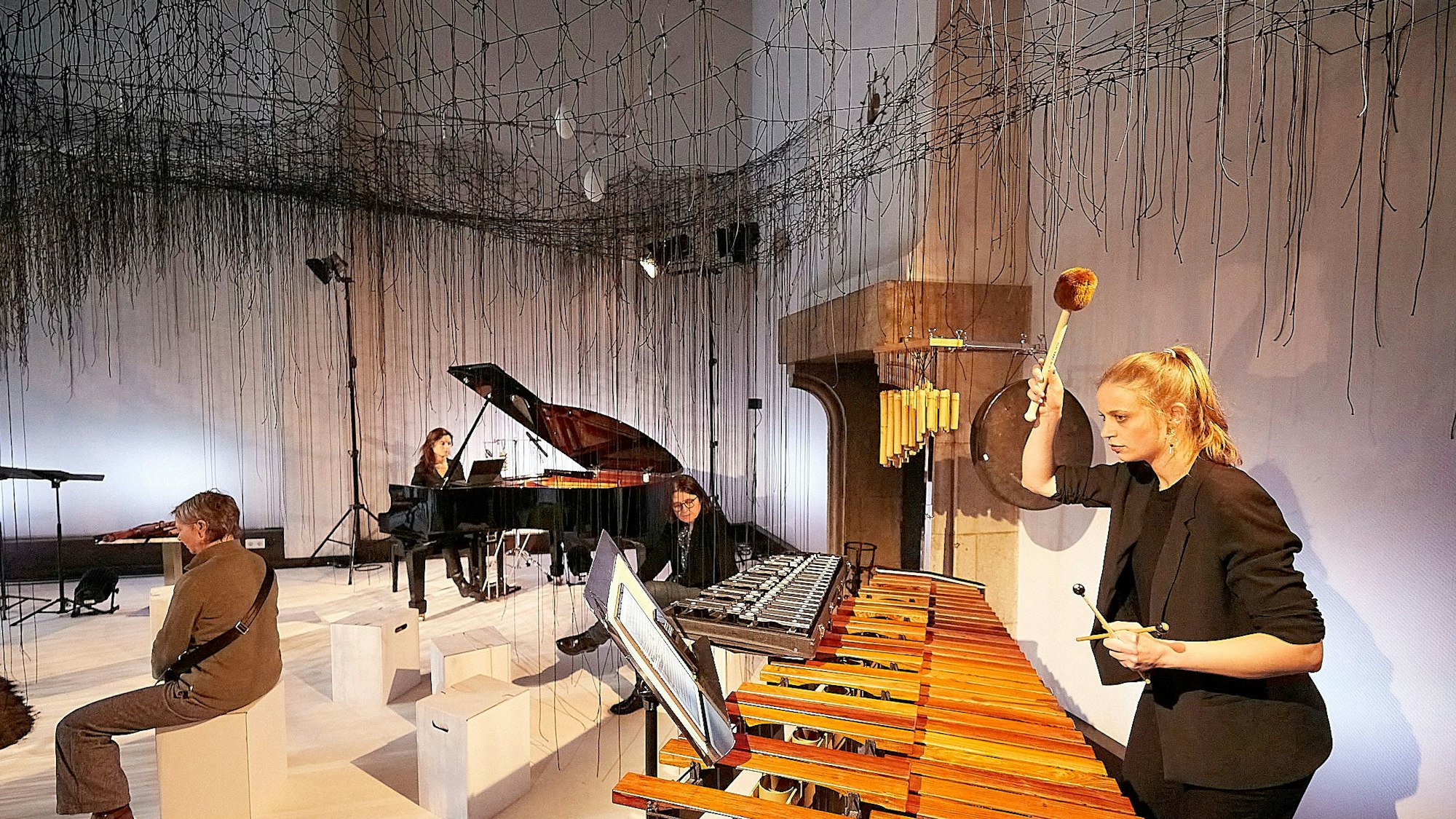 Die Musikerinnen spielen in einem Raum, von dessen Decke Tausende von Fäden herabhängen.