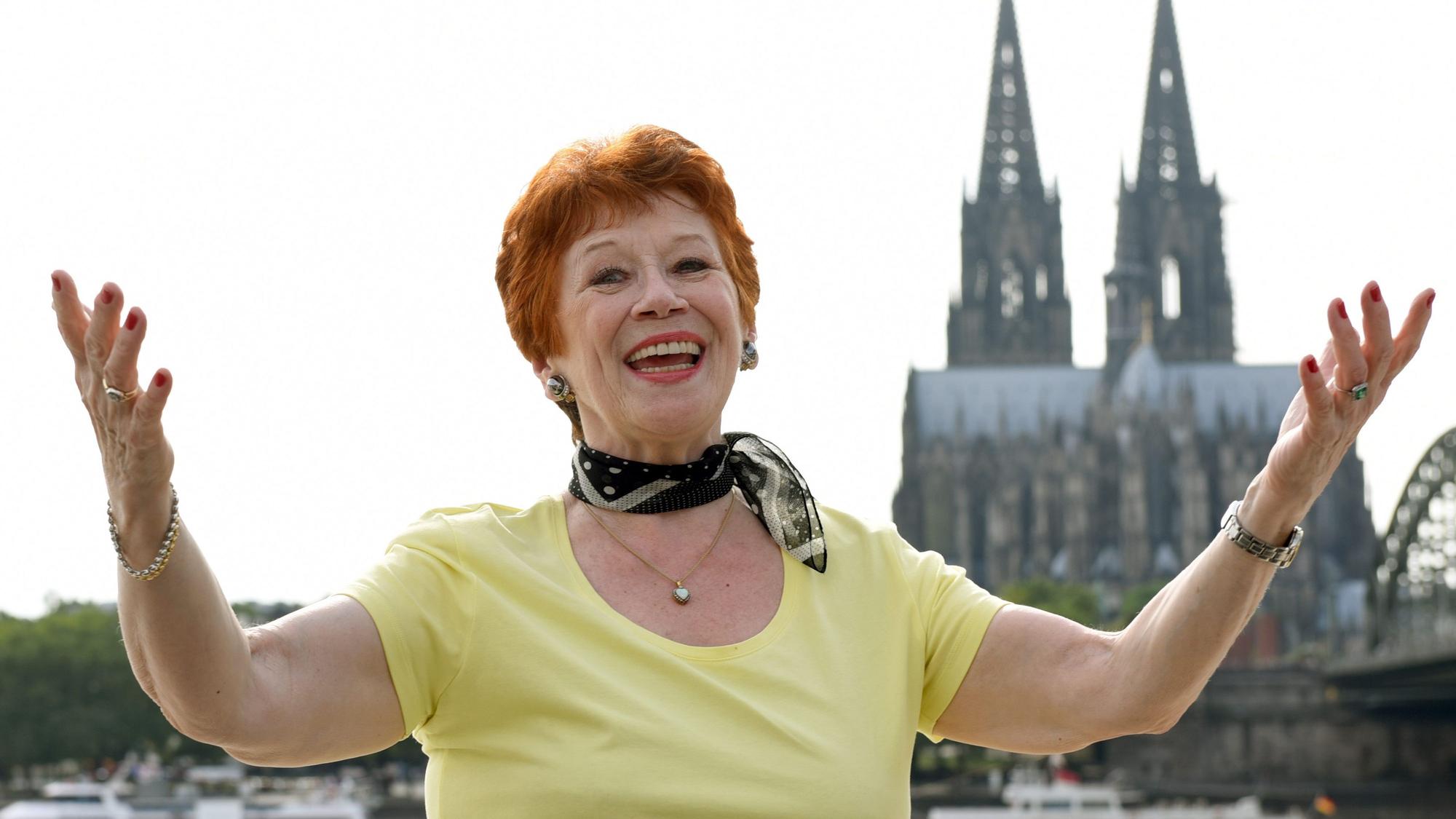 Die Schauspielerin Lotti Krekel steht bei einem Foto-Shooting anlässlich ihres 75. Geburtstags mit ausgebreiteten Armen am Rheinufer vor der Kulisse des Doms.