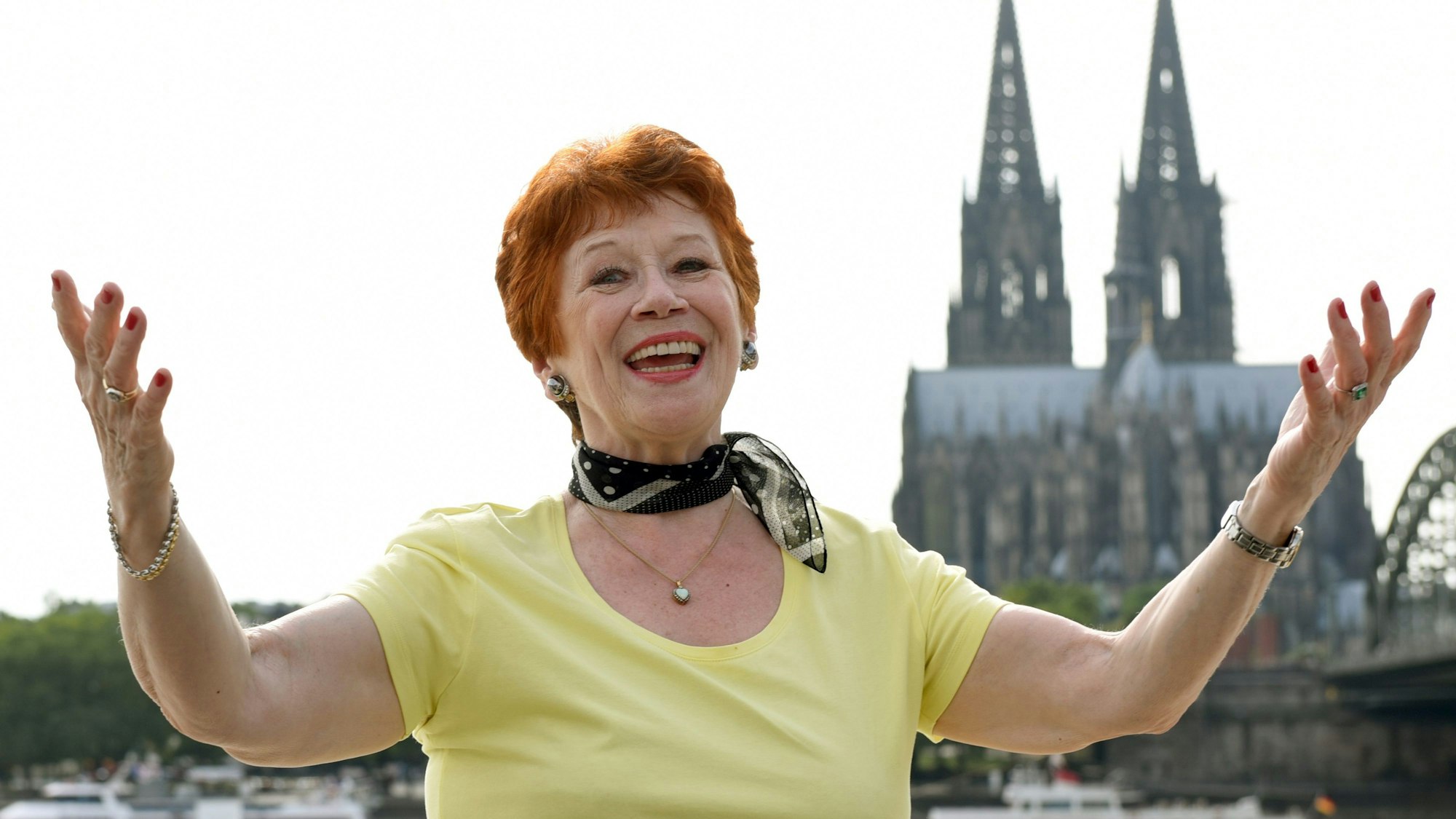 Die Schauspielerin Lotti Krekel steht bei einem Foto-Shooting anlässlich ihres 75. Geburtstags mit ausgebreiteten Armen am Rheinufer vor der Kulisse des Doms.