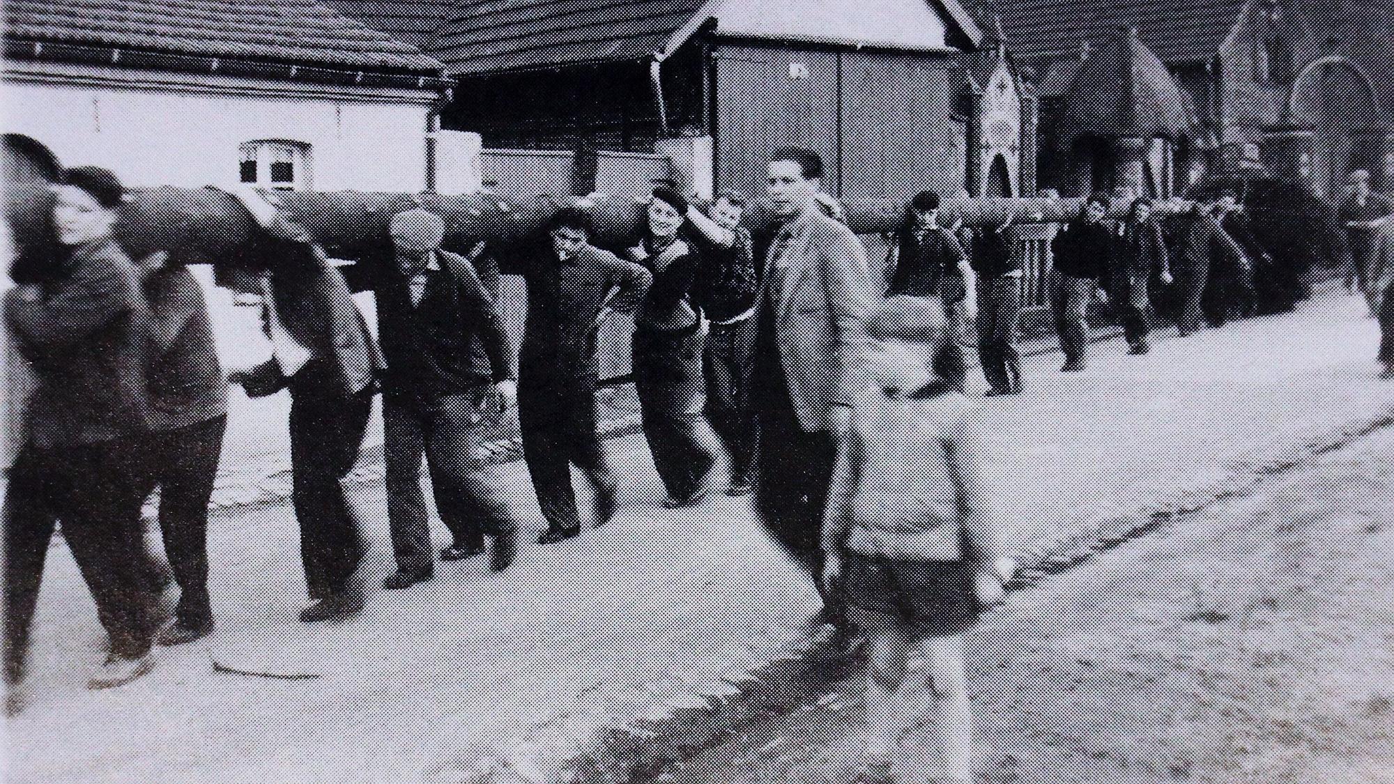 Es sind mehrere Männer zu sehen, die einen riesigen Baumstamm tragen. Das Bild ist schwarz-weiß.
