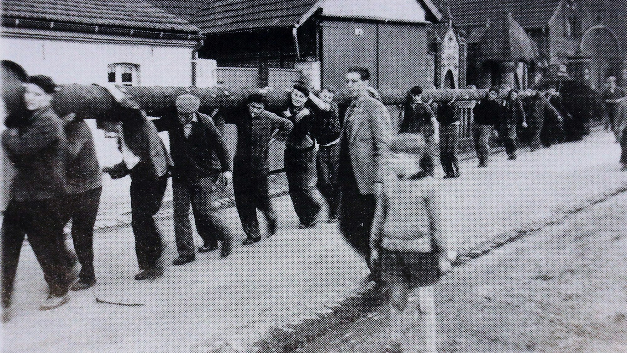 Es sind mehrere Männer zu sehen, die einen riesigen Baumstamm tragen. Das Bild ist schwarz-weiß.