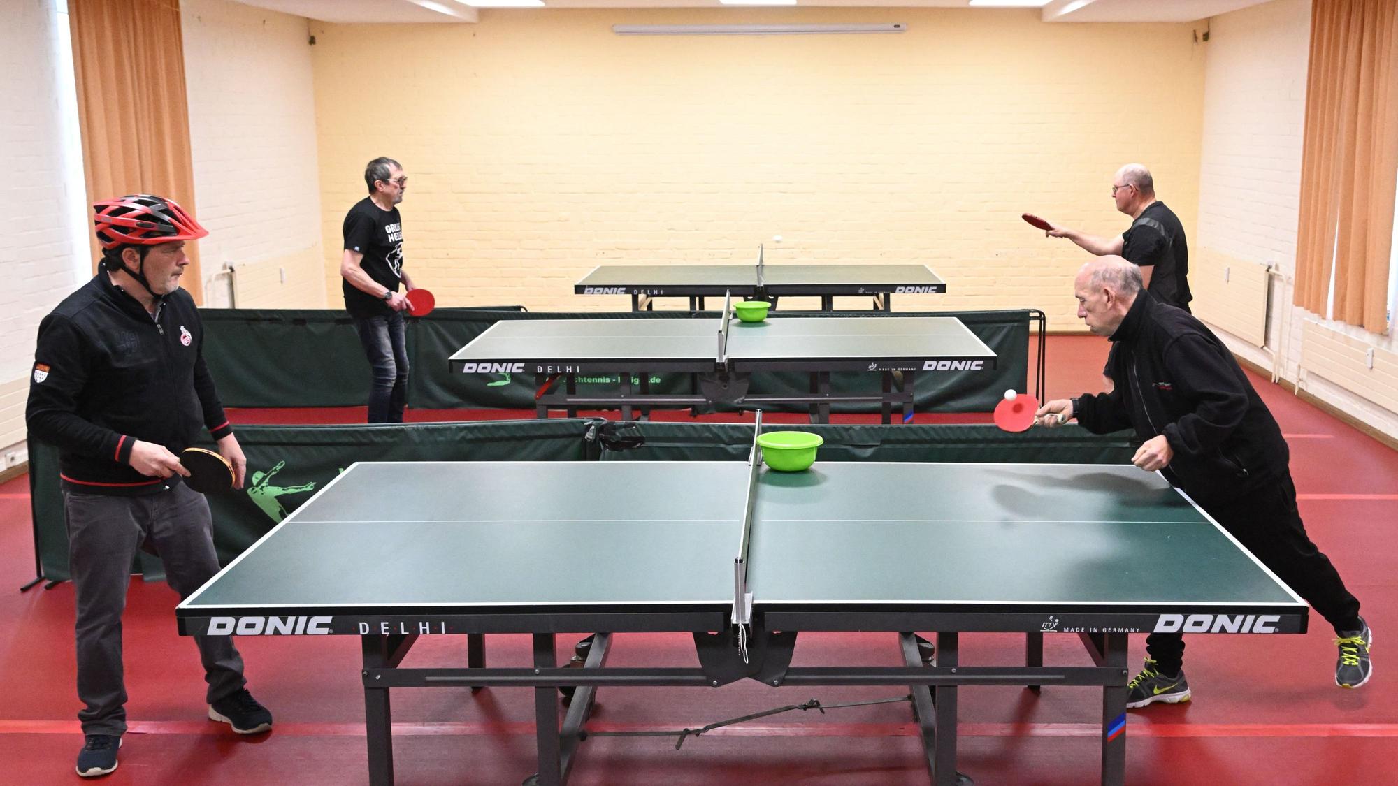 Drei Tischtennisplatten auf rotem Boden, vier ältere Herren in schwarzer Kleidung spielen an zwei Platten Tischtennis.