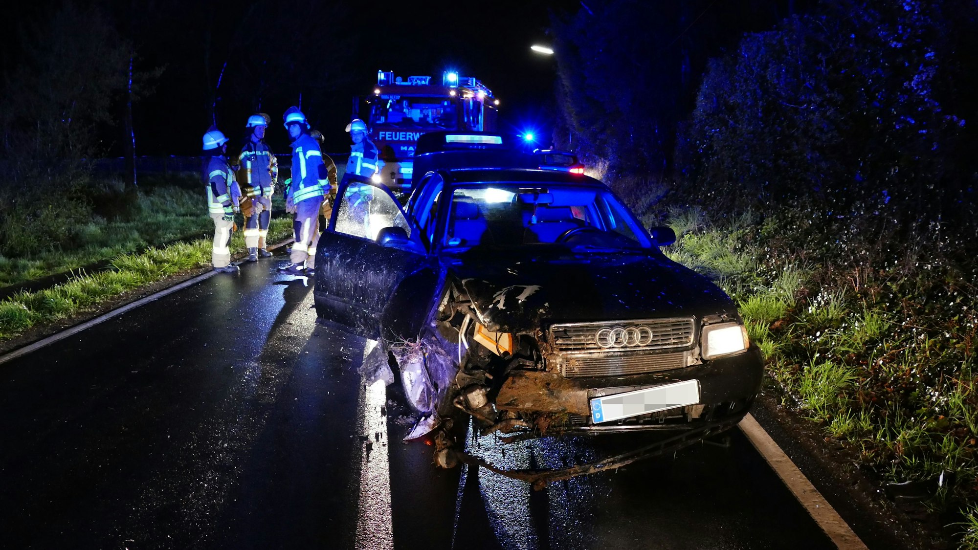 Ein Wagen steht demoliert auf einer Straße. Es ist dunkel. Feuerwehrleute stehen dahinter.