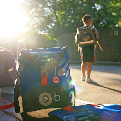 Schulranzen stehen am Morgen auf einem Schulhof. Ein Junge mit Schulranzen steht davor.