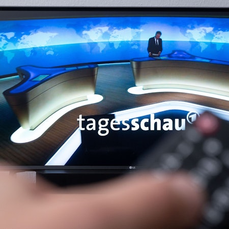 Ein Mann hält eine Fernbedienung vor einen Fernseher, auf dem die Tagesschau läuft.