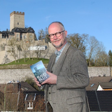 Krimiautor Ralf Kramp steht vor der Burg Kerpen, die auch auf dem Cover seines neuen Buches zu sehen ist, das er in der Hand hält.
