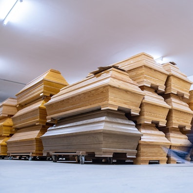 Mehrere Särge aus Holz stehen übereinander gestapelt in einem Raum. Ein Mann geht an ihnen vorbei.