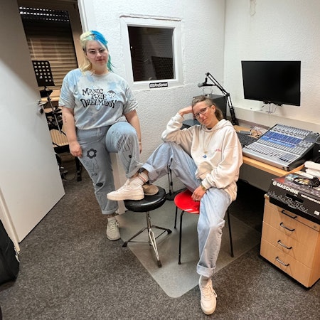 Zwei junge Frauen, sportlich gekleidet, sitzen lässig und mit jeweils einem Fuß auf einen Stuhl gestellt, in einem Tonstudio.