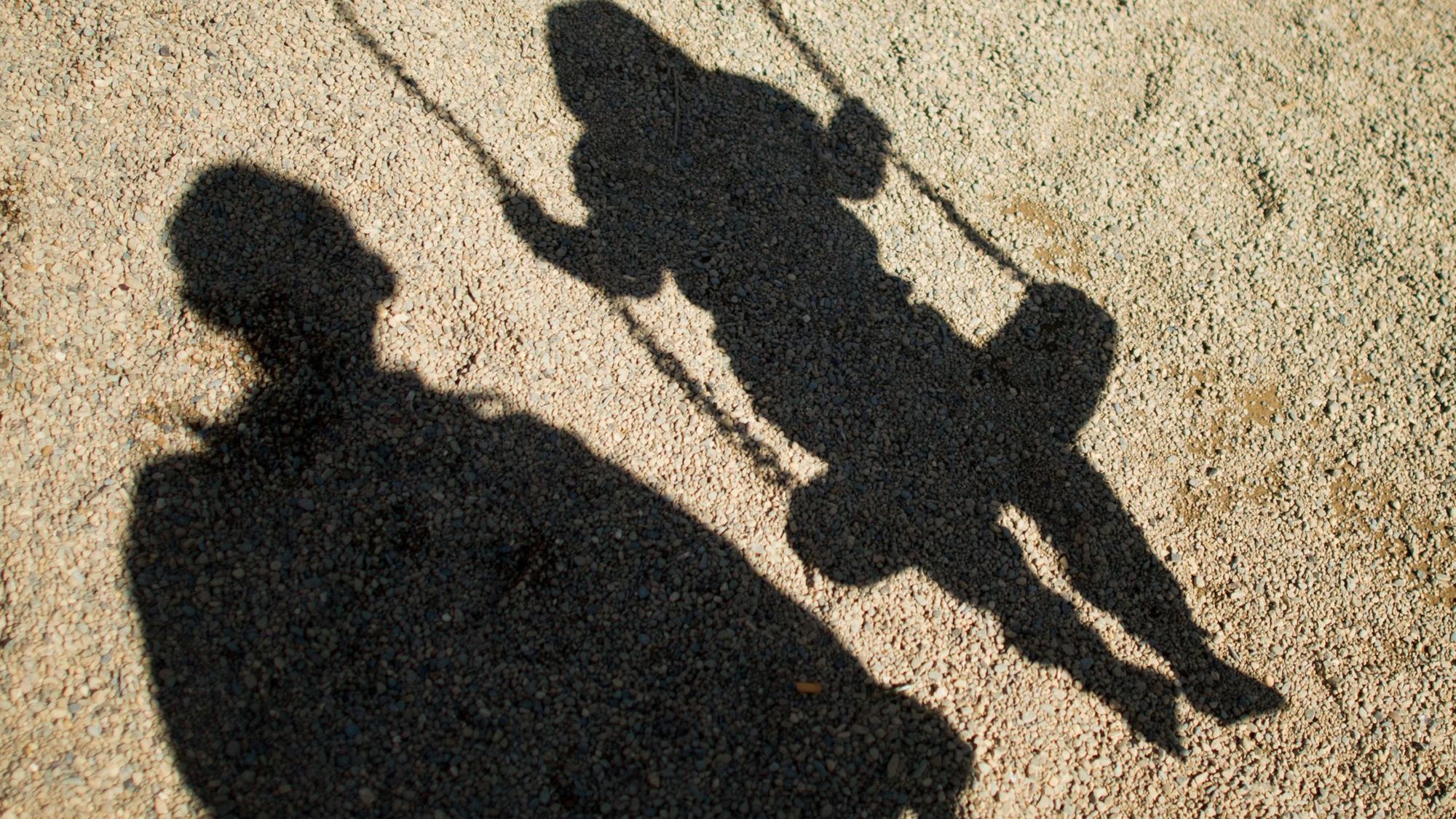 Der Schatten von einem Mann und einem schaukelnden Kind fallen auf Sand auf einem Spielplatz.
