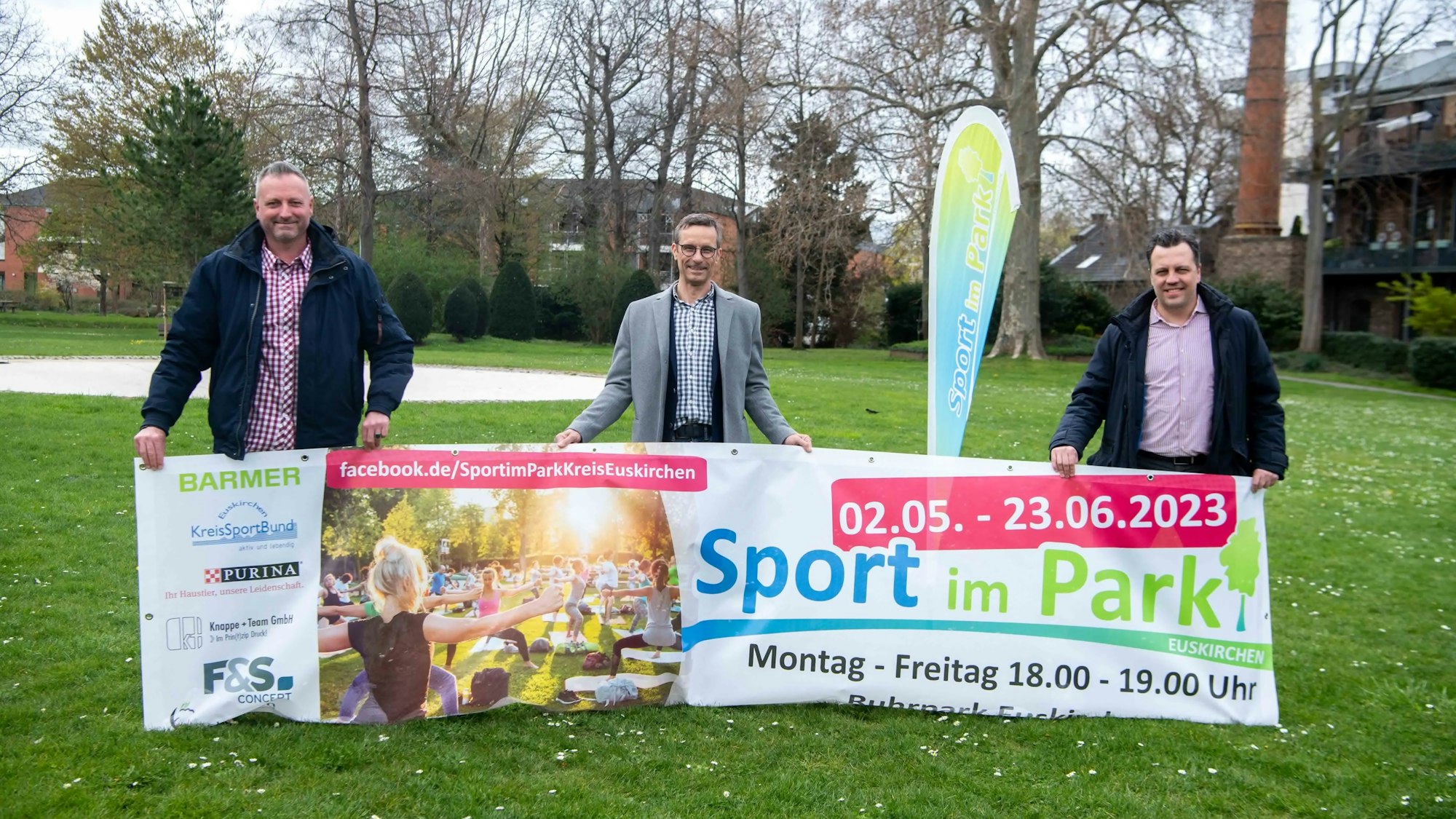 Die drei Männer posieren mit dem Banner von „Sport im Park“ und lächeln in die Kamera.