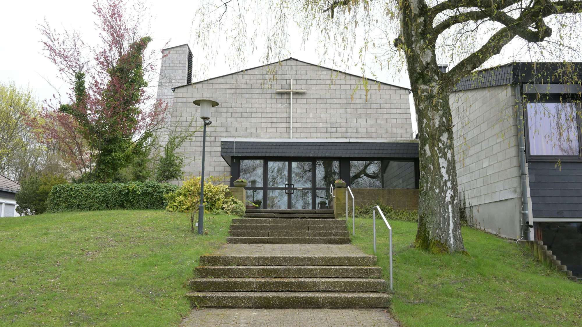 Vor dem Kirchengebäude mit Glockenturm und Kreuz über dem Eingang steht rechts ein Baum.