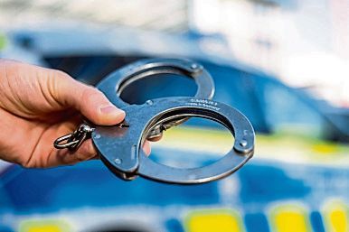 Das Foto zeigt Handschellen, im Hintergrund ist verschwommen ein Polizeiwagen zu sehen.