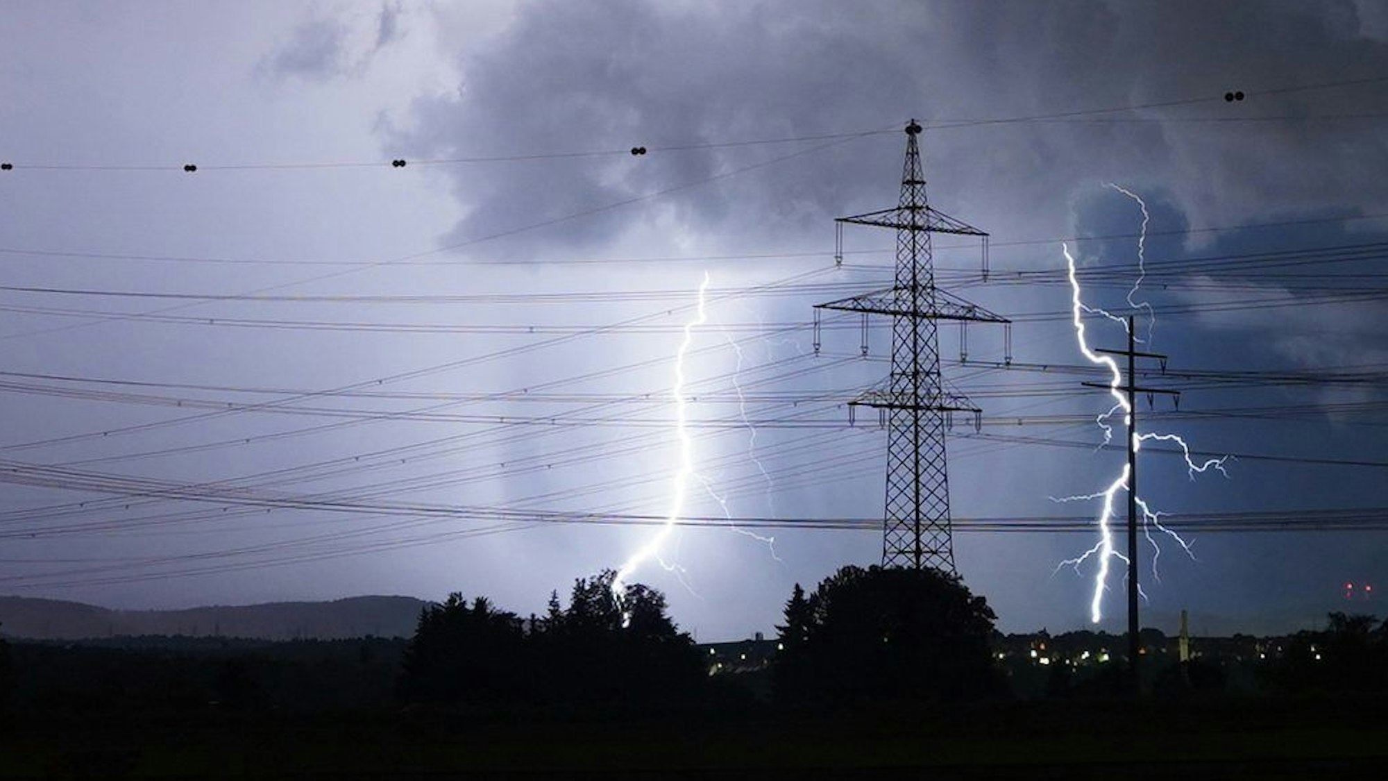 Zwei Blitze schlagen in der Nähe eines Strommastes ein, der Himmel ist stark verdunkelt. Im Hintergrund sind einige Häuser zu sehen. (Symbolbild)
