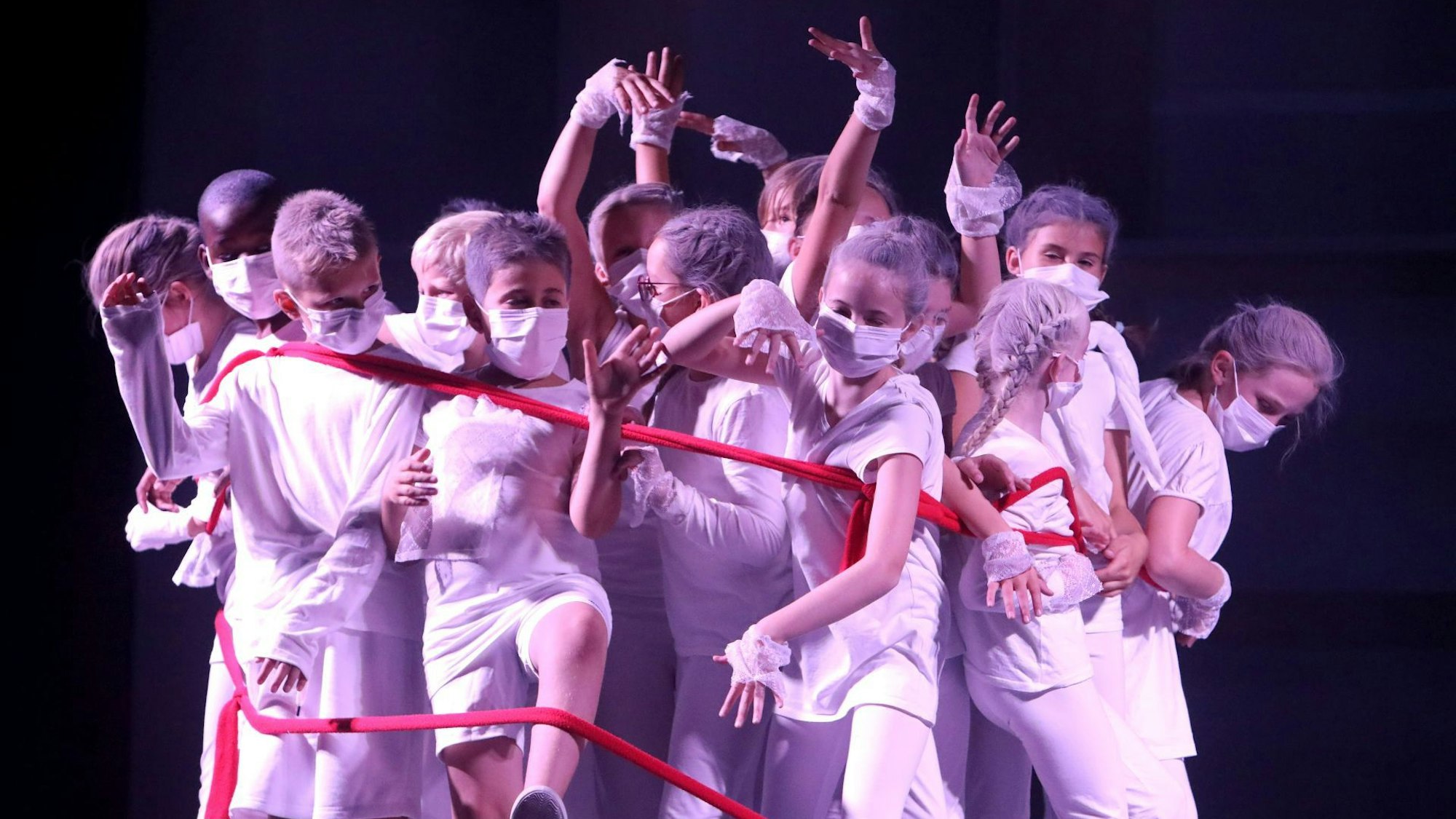 Eine Gruppe junger Menschen mit Mundschutz tanzt in lilafarbenem Licht, verbunden durch ein rotes Seil.