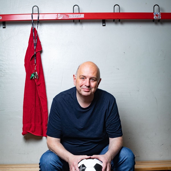 Frank Goosen sitzt auf einer Bank, in einem blauen T-Shirt vor einer weißen Wand. Links von ihm hängt ein rotes Handtuch und ein Schlüsselbund mit Trillerpfeife.